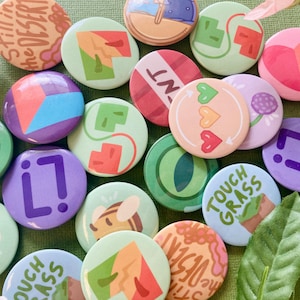May include: A selection of colourful round pinback buttons with diverse designs. Some buttons feature geometric shapes, while others display text like "Touch Grass" and "Still in the Desert." The buttons are in shades of green, purple, pink, and orange.