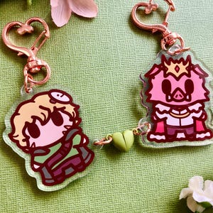 May include: Two character charms on keychains. One charm is a pink pig wearing a crown and a white and red robe. The other charm is a green and brown character with blonde hair and a white headband. The charms are connected by a green heart charm.