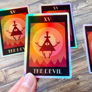 Puede incluir: Pegatinas holográficas que representan un personaje triangular rojo con un ojo, brazos y piernas, sobre un fondo degradado de naranja, amarillo y rojo. El texto "THE DEVIL" está en la parte inferior, con "XV" en la parte superior.