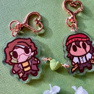 Puede incluir: Dos llaveros acrílicos transparentes con diseños de personajes de dibujos animados. Uno presenta un personaje con cabello castaño, un parche en el ojo y un abrigo marrón. El otro tiene un personaje con gorra roja y camisa blanca. Ambos tienen cierres en forma de corazón.