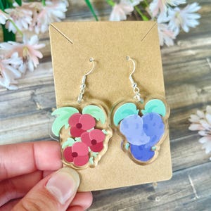 以下が含まれることがあります： 花柄のデザインのアクリルイヤリングのペア。片方のイヤリングは緑の葉を持つ赤い花、もう片方は緑の葉を持つ青い花です。イヤリングは銀色のフックに取り付けられています。