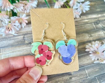 May include: A pair of acrylic earrings with a floral design. One earring features red flowers with green leaves, and the other has blue flowers with green leaves. The earrings are attached to silver-coloured hooks.