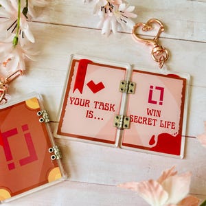 May include: Clear acrylic keychains with a book design and the text "YOUR TASK IS..." and "WIN SECRET LIFE". The keychains have a red and brown color scheme, with gold-colored hardware and a heart-shaped clasp.