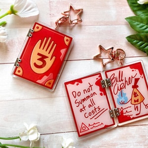 May include: Red and clear acrylic keychains featuring designs from a cartoon series. One keychain is a red book with a hand and the number 3. The other keychain is a book that opens to reveal the text "Do not summon at all costs" and a character illustration.