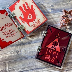 May include: Clear acrylic book-shaped charms with red and black graphics. One charm has the text "TO SUMMON: Place your hand on the next page." Another has a red handprint and the text "time to get WEIRD." A third charm features a triangle with an eye and the name "Bill."