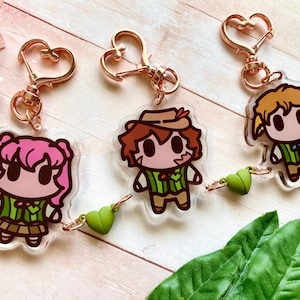 May include: Three character keyrings with rose gold heart-shaped clasps. Each keyring features a different cartoon character with brown, pink, and blonde hair, wearing green and brown outfits. Small green heart charms dangle below.