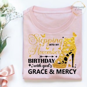 May include: Pink t-shirt with a gold glitter high heel shoe and the text "Stepping into my November Birthday with God's Grace & Mercy".