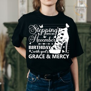 May include: Black t-shirt with white text that reads "Stepping into my November Birthday with God's Grace & Mercy". A white high-heeled shoe with straps is featured on the shirt.