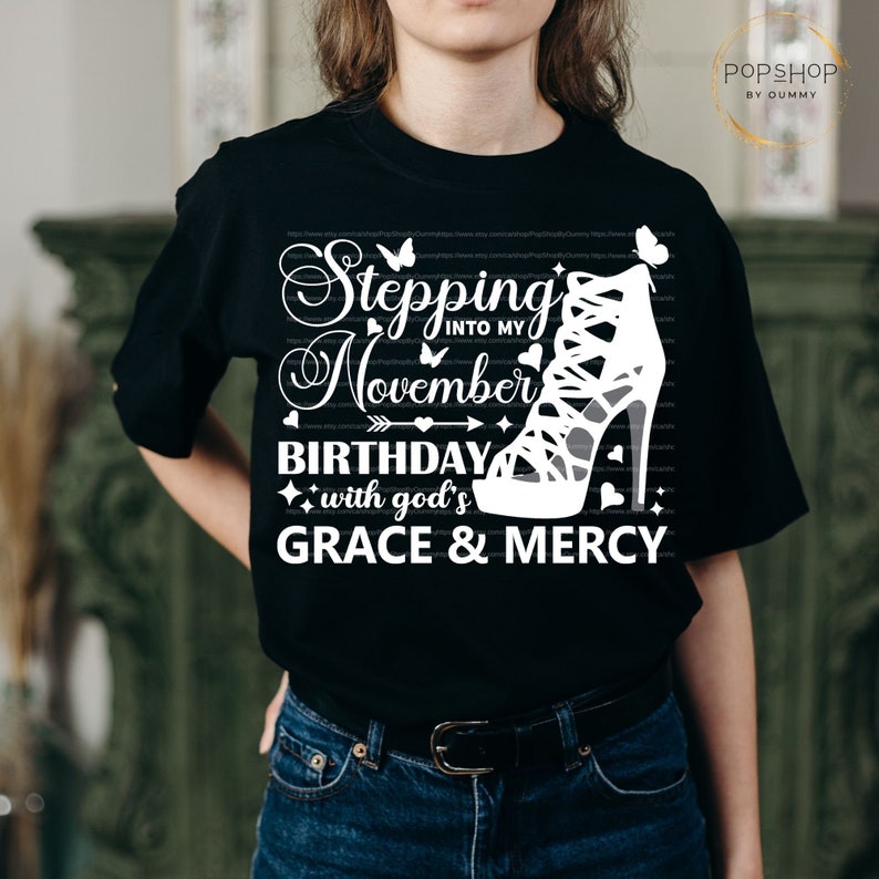 May include: Black t-shirt with white text that reads "Stepping into my November Birthday with God's Grace & Mercy". A white high-heeled shoe with straps is featured on the shirt.
