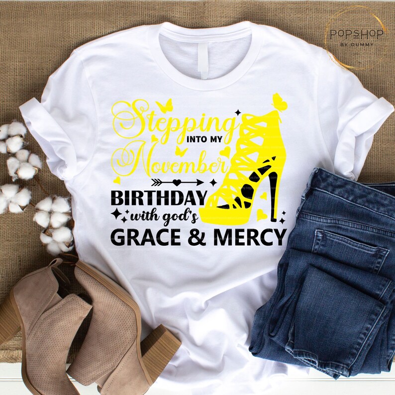 May include: White t-shirt with black text that reads "Stepping into my November Birthday with god's GRACE & MERCY". A yellow high-heeled shoe with a butterfly on the side is in the text.