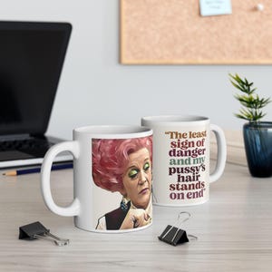 May include: Two white ceramic mugs. One mug features a portrait of a woman with pink hair and green eyeshadow. The other mug has the quote, "The least sign of danger and my pussy's hair stands on end."