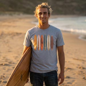 May include: A gray t-shirt featuring a graphic of six surfboards in various colors. The surfboards are vertically aligned. The person in the image is holding a wooden surfboard on a beach.