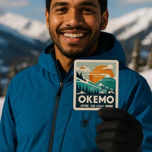 May include: A sticker featuring a mountain landscape with the word "OKEMO" in white text. The sticker also includes the text "AUTITUDE LT3H-3 31411 VERMONT". The sticker is held by a person wearing a blue jacket and black gloves.