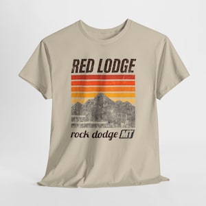 以下が含まれることがあります： ベージュのTシャツ。デザインには、赤、オレンジ、黄色の水平ストライプの上に「RED LODGE」の文字が含まれています。ストライプの下には山脈のグラフィックがあり、「rock dodge MT」の文字があります。
