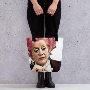 May include: White tote bag featuring a stylized portrait of a woman with pink hair and green eyeshadow. The tote bag has black straps and is held by a person wearing black pants and boots.