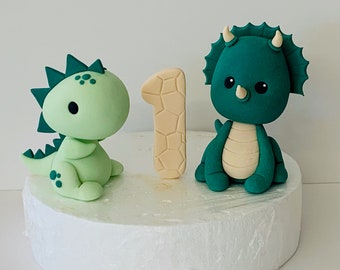 May include: Green and beige fondant dinosaur cake toppers, including a triceratops and a smaller dinosaur, with a beige number one in the center.