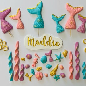 Op de afbeelding: Een set met zeemeermin-thema caketoppers, waaronder een zeemeerminstaart, schelpen en spiralen. De toppers zijn in tinten roze, blauw en goud. De set bevat ook een caketopper met de naam "Madolvie" geschreven in gouden letters.