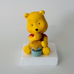 Könnte beinhalten: Eine gelbe Winnie Puuh-Figur, die auf einem weißen Kuchensockel sitzt. Pooh hält einen Honigtopf, auf dem "Hunny" steht.