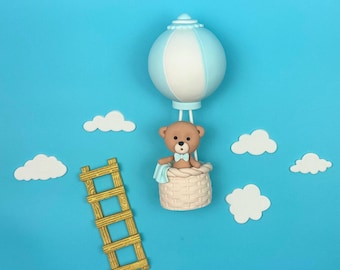 May include: A whimsical scene featuring a teddy bear in a hot air balloon, with a light blue and white balloon, a woven basket, and a small wooden ladder. White cloud cutouts are scattered on the bright blue background.