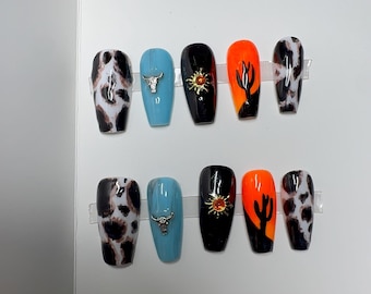 May include: A set of ten artificial nails with a western theme. Designs include a cow print, a silver bull head, a black nail with a gold sun, and an orange nail with a black cactus. The nails are in a coffin shape.