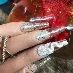 May include: Long, clear, acrylic nails with a gold glitter design. Some nails have a white 3D flower design.