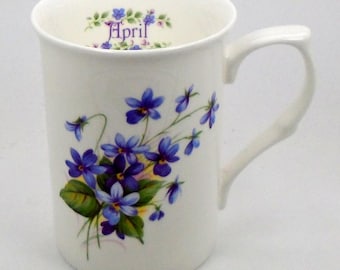 May include: A white ceramic mug with a floral design featuring blue and purple violets. The mug has the word "April" printed on the inside rim.