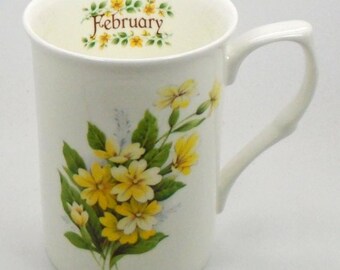 May include: White ceramic mug with a floral design featuring yellow primroses and green leaves. The mug has the word "February" written in brown lettering on the inside rim.