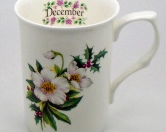 May include: White ceramic mug with a floral design featuring white flowers and green leaves. The mug has the word "December" written on the inside rim.