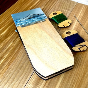 May include: A wooden weaving tool with a metal comb, green and blue thread spools, and a needle. The tool is rectangular with a light wood finish and black trim. The spools are wooden with heart-shaped cutouts.