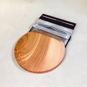 Könnte beinhalten: Ein hölzernes Kalimba mit einem runden, hellbraunen Resonanzkörper und Metallzungen. Das Instrument ist teilweise sichtbar, wobei die Zungen über der Holzbasis aufgefächert sind. Das Kalimba ist ein Musikinstrument, das oft zur Entspannung verwendet wird.