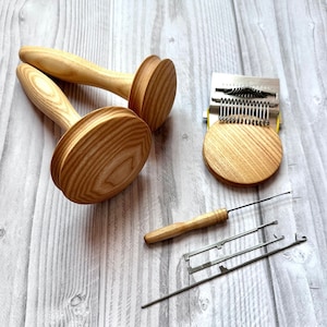 May include: Two wooden darning mushrooms with light wood grain, a metal darning loom tool with a circular wooden base, a small wooden-handled needle, and two thin metal darning needles arranged on a light-coloured wood-look surface.
