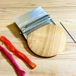 Op de afbeelding: Een weefgereedschapsset met een metalen frame, houten basis en een houten naald. Er zijn ook twee bundels kleurrijk borduurgaren in roze en oranje tinten. De set staat op een licht houten oppervlak.