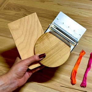 Könnte beinhalten: Ein hölzerner Webstuhl mit rechteckiger Holzbasis und rundem Holzaufsatz. Der Webstuhl hat Metallkomponenten und wird von orangefarbenem und pinkfarbenem Garn und einer Nadel begleitet. Das Holz hat eine hellbraune Farbe.