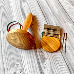 May include: A wooden darning mushroom with a smooth handle and a rounded top, accompanied by a small metal weaving loom tool with a circular wooden base and two thin metal prongs, all featuring natural wood grain patterns and green elastic bands.