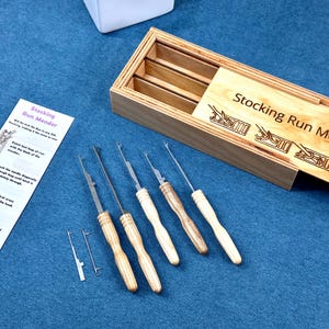 May include: A wooden box set with a sliding lid, labelled "Stocking Run Mender," contains several mending tools with wooden handles and metal needles. An instruction card is also included, all on a blue surface.