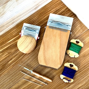 May include: A collection of wooden weaving tools and spools of thread. The set includes two wooden weaving looms, a needle, and two spools of green and blue thread. The items are arranged on a wooden surface.