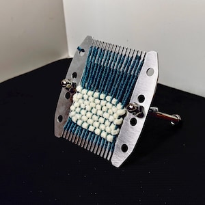 May include: A metal darning loom tool featuring a silver-colored frame with multiple teeth and circular cutouts. The device holds a woven fabric patch created with dark blue vertical warp threads and a central section of thick, cream-colored horizontal weft.