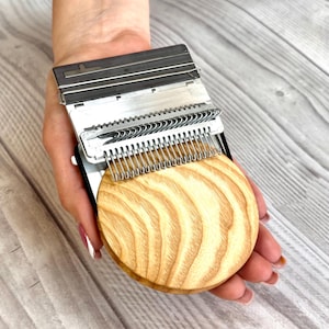 Könnte beinhalten: Ein silbernes und schwarzes Webwerkzeug mit Holzsockel. Das Werkzeug hat eine Reihe von Metallzähnen und eine rechteckige Oberseite. Der Holzsockel ist rund mit einem hellbraunen Holzmaserungsmuster.