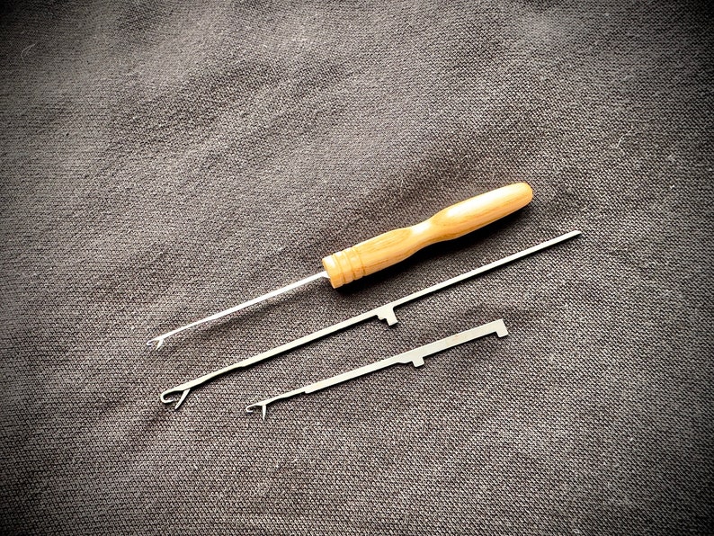 May include: Three crochet hook tools on a dark gray fabric background. One tool has a wooden handle and two tools have metal handles.