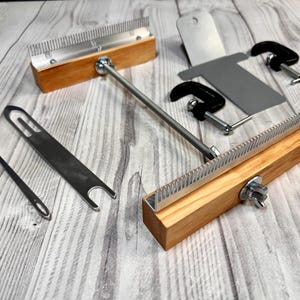 May include: A set of weaving tools including two wooden-handled metal combs connected by a metal rod, a long metal needle, a flat metal tool with a slotted end, a metal plate, and two small black metal C-clamps for securing the equipment.