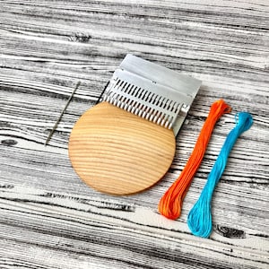 Op de afbeelding: Een weefset met een houten basis, metalen kam en naald. Twee strengen borduurgaren in oranje en blauw liggen naast het weefgereedschap. De items liggen op een houtnerf oppervlak.