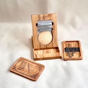 May include: A wooden darning loom tool with a metal weaving head and a round wooden base, accompanied by a small rectangular wooden box containing metal darning needles and a separate wooden lid engraved with the text DARNING NEEDLES.