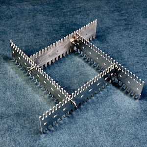 May include: A silver-colored metal frame constructed from four interlocking notched strips. Each strip features a series of evenly spaced teeth along the top edge and small circular holes along the body, connected by metal fasteners at the corners.