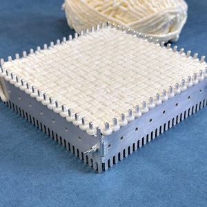 May include: A square metal weaving loom featuring silver-coloured vertical pegs along the top edges and a slotted base. The loom is currently holding a woven square of thick, off-white yarn, with a ball of the same yarn resting behind the frame.