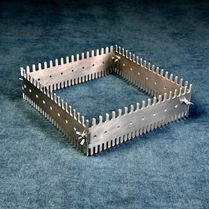 May include: A square metal frame featuring a series of evenly spaced vertical prongs along the top edge and small circular holes along the side. The frame is constructed from four interlocking panels secured at the corners with metal wing nuts.
