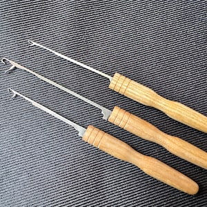 以下が含まれることがあります： 木製のハンドルと金属製の先端を持つクロシェフックが3つ。フックはサイズが異なり、暗いグレーのテクスチャのある表面に並べて配置されています。