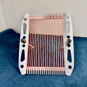 Op de afbeelding: Een zilverkleurig metalen stopraam met een rechthoekig frame en gekartelde randen aan de boven- en onderkant. Het raam is bespannen met verticale draden in perzikkleur, vastgezet met twee kleine metalen bouten op de zijpanelen.
