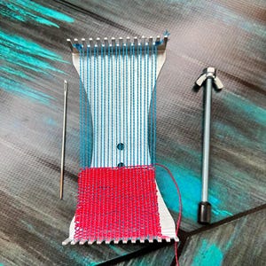Può includere: Un telaio in metallo con fili blu e tessuto rosso intrecciato. Sono presenti anche un ago e una vite. Il telaio è su una superficie a motivi.