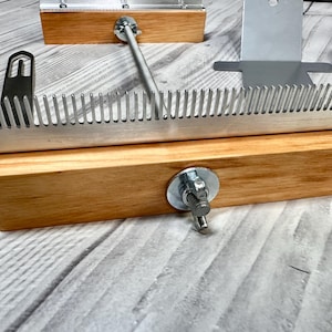 May include: A weaving comb tool featuring a long, silver-coloured metal comb with fine teeth attached to a rectangular light-brown wooden base. The base includes a central metal bolt with a wing nut, and two separate metal needles are nearby.