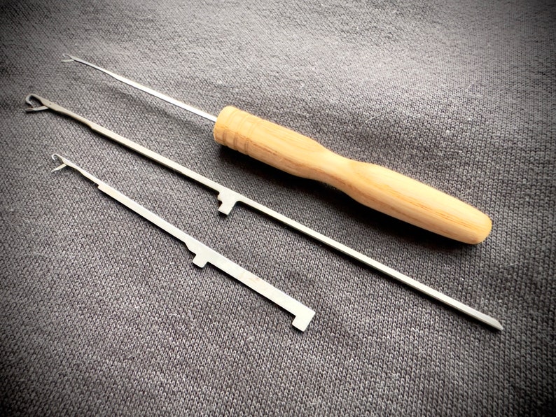 May include: Three knitting tools, two with metal hooks and one with a wooden handle. The tools are laid out on a gray fabric surface.